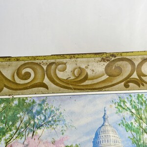 K&ouml;nnte beinhalten: Vintage Blechdose mit goldfarbenem Rand und einem dekorativen Schn&ouml;rkeldesign. Die Dose zeigt ein Aquarellbild des US-Kapitols mit B&auml;umen im Vordergrund.