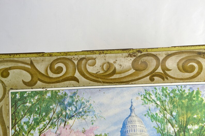 K&ouml;nnte beinhalten: Vintage Blechdose mit goldfarbenem Rand und einem dekorativen Schn&ouml;rkeldesign. Die Dose zeigt ein Aquarellbild des US-Kapitols mit B&auml;umen im Vordergrund.