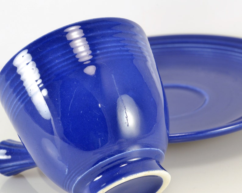 May include: A blue ceramic teacup and saucer set. The cup has a rounded shape and a handle. The saucer is round and has a slightly raised edge.