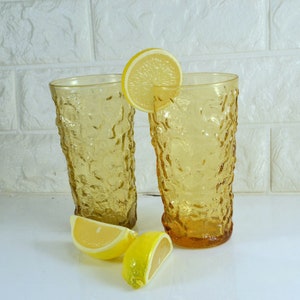 May include: Two amber glass tumblers with a textured surface, each with a lemon wedge on top. The tumblers are sitting on a white surface.