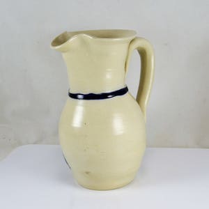 May include: A white ceramic pitcher with a blue band around the middle and a handle. The pitcher has a slightly rounded shape.