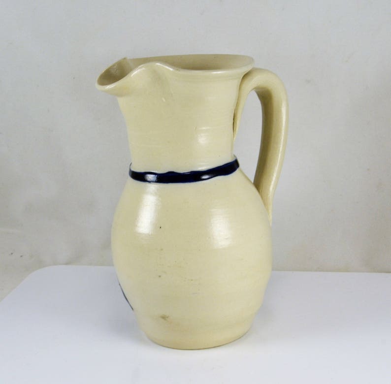 May include: A white ceramic pitcher with a blue band around the middle and a handle. The pitcher has a slightly rounded shape.