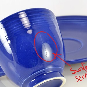 May include: A blue ceramic teacup and saucer set with a surface scratch. The cup has a handle and a rounded shape. The saucer is round and has a slight lip.