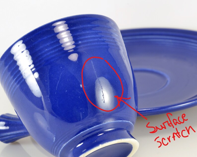 May include: A blue ceramic teacup and saucer set with a surface scratch. The cup has a handle and a rounded shape. The saucer is round and has a slight lip.