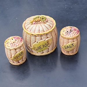 May include: A ceramic kitchen set with a basket weave design. The set includes a lidded container with the word "GREASE" on a leaf, and two smaller shakers labeled "PEPPER" and "SALT". The lids are decorated with grapes.