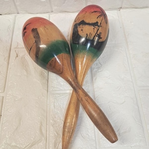 May include: Two wooden maracas featuring a hand-painted tropical sunset scene with palm trees and silhouettes in shades of orange, green, and black. The instruments have a smooth, polished finish and are crossed over each other in the center.