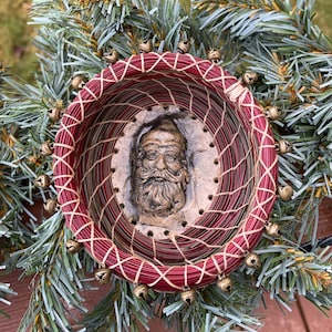 May include: A red and green woven basket with a Santa Claus face in the center. The basket is decorated with small gold bells around the rim.