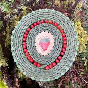 May include: A handcrafted, circular basket with a green and natural fiber weave. The basket features a central ceramic strawberry design, surrounded by red berry-like beads and green glass beads. The basket is set against a natural background.