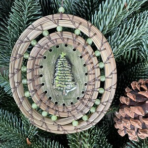 May include: A round, woven ornament with a green tree design in the center. The ornament is decorated with green beads and has a brown string for hanging.