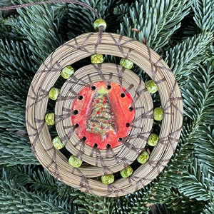 May include: A round, woven ornament with a brown cord and green beads. The center of the ornament features a red and green ceramic tile with a Christmas tree design.