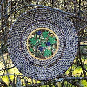 May include: A circular woven wall hanging with a purple and beige design. The center of the hanging features a cutout of a vine with green leaves and black grapes.