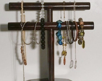May include: A dark brown wooden jewelry stand with two horizontal bars for hanging necklaces and bracelets. The stand has a hexagonal base and features a variety of necklaces and bracelets in different colors and styles.