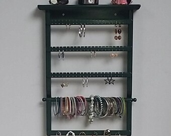 Puede incluir: Un organizador de joyas de madera verde oscuro con múltiples filas de ganchos para pendientes, una barra para pulseras y ganchos para collares.