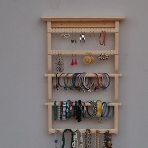 Op de afbeelding: Een houten sieradenorganisator met meerdere rijen om oorbellen, armbanden en kettingen op te hangen. De organisator heeft een lichte houtkleur en is aan een muur gemonteerd.