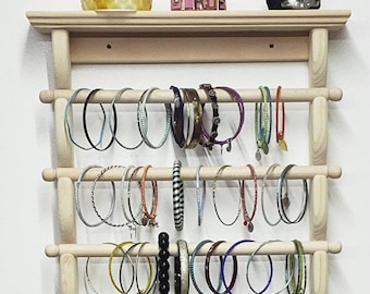 Puede incluir: Un organizador de joyas de pared de madera con múltiples clavijas para colgar pulseras y collares. El organizador tiene un estante superior para guardar otros artículos. El organizador está hecho de madera natural y tiene un aspecto rústico.