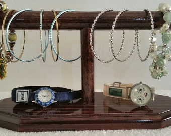 May include: A wooden jewelry stand with a brown finish. The stand has two levels, one for bracelets and one for watches. There are several bracelets on the top level, including a beaded bracelet with green and yellow beads, a silver chain bracelet, and a bracelet with green and white beads. There are two watches on the bottom level, one with a blue band and one with a silver band.