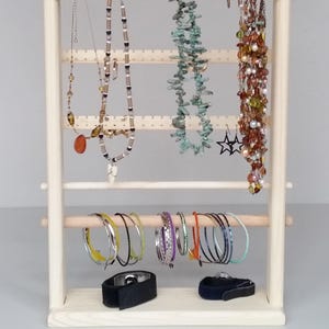 May include: A wooden jewellery stand with multiple tiers for hanging necklaces and earrings. The stand also has a horizontal bar for hanging bracelets. There are several necklaces, earrings, and bracelets on the stand.