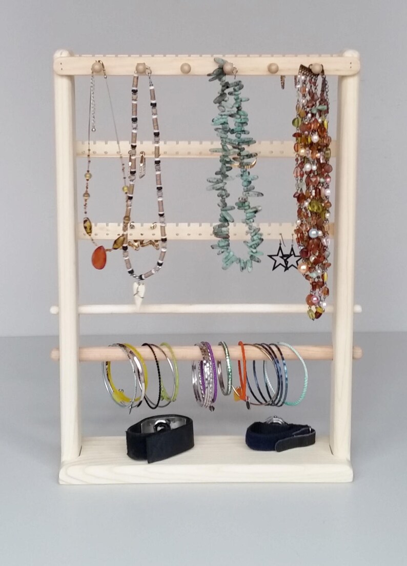 May include: A wooden jewellery stand with multiple tiers for hanging necklaces and earrings. The stand also has a horizontal bar for hanging bracelets. There are several necklaces, earrings, and bracelets on the stand.