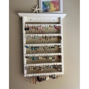 May include: A white wooden wall-mounted earring organizer with six horizontal slats featuring numerous hanging holes. The rack holds a variety of colorful dangling earrings. A small white starfish and a framed rainbow print sit on the top shelf.
