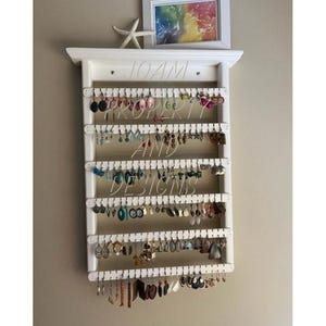 May include: A white wooden wall-mounted earring organiser with six horizontal slats featuring numerous hanging holes. The rack holds a variety of colourful dangling earrings. A small white starfish and a framed rainbow print sit on the top shelf.