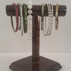 May include: A wooden bangle stand with a square base and a T-shaped top. The stand is dark brown and has a smooth finish. There are several bangles on the stand, including silver, gold, green, and pink.