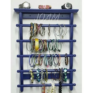 Op de afbeelding: Een donkerblauwe houten wandorganizer voor sieraden met vijf horizontale stangen voor het ophangen van armbanden, met een bovenplank waarop twee polshorloges en een klein decoratief blokje met het woord TRUE staan.