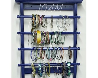 Puede incluir: Un organizador de joyas de pared en madera azul oscuro con cinco barras horizontales para colgar pulseras, con un estante superior que sostiene dos relojes de pulsera y un pequeño bloque decorativo con la palabra TRUE impresa.