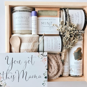 May include: A wooden gift box filled with self-care items, including a sugar scrub, soap, body butter, and tea. The box also includes a small wooden spoon, a washcloth, and a card that says "You got this Mama".