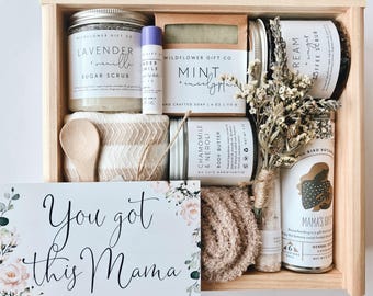 Może przedstawiać: Drewniana skrzynka prezentowa wypełniona produktami do pielęgnacji, w tym peelingiem cukrowym, mydłem, masłem do ciała i herbatą. Skrzynka zawiera również małą drewnianą łyżkę, ściereczkę i kartkę z napisem "You got this Mama".