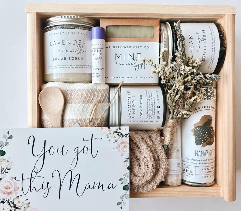 May include: A wooden gift box filled with self-care items, including a sugar scrub, soap, body butter, and tea. The box also includes a small wooden spoon, a washcloth, and a card that says "You got this Mama".