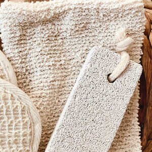 May include: A spa set featuring a textured, off-white washcloth, a pumice stone with a rope handle, and a round, woven sponge. The items are arranged on a woven tray, creating a natural, spa-like aesthetic.