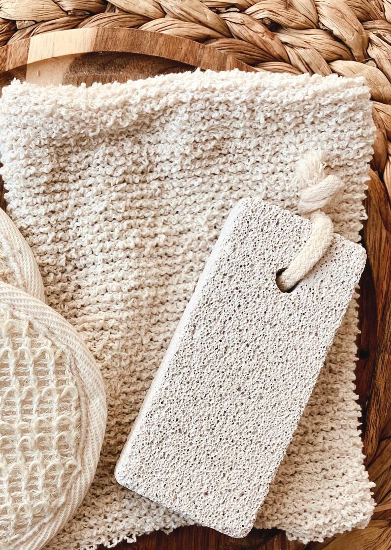 May include: A spa set featuring a textured, off-white washcloth, a pumice stone with a rope handle, and a round, woven sponge. The items are arranged on a woven tray, creating a natural, spa-like aesthetic.