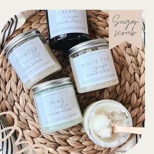 May include: Four glass jars with silver lids filled with sugar scrubs. The jars are labeled with the names of the scents: Lavender Vanilla, Mint + Cedar, White Tea + Ginger, and Cream + Sugar Coffee. The jars are arranged on a woven wicker surface.