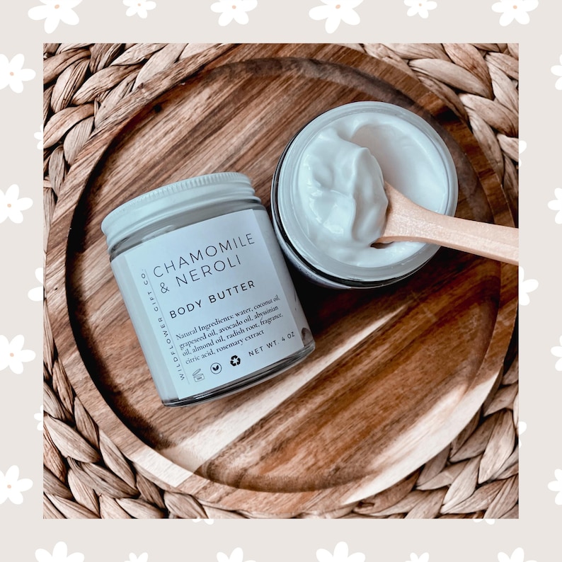 May include: A white jar of chamomile and neroli body butter with a white lid. The jar is on a wooden surface with a wooden spoon scooping out some of the butter. The jar has a label that says "Wildflower Gift Co. Chamomile & Neroli Body Butter" and lists the ingredients. The jar is 4 oz.