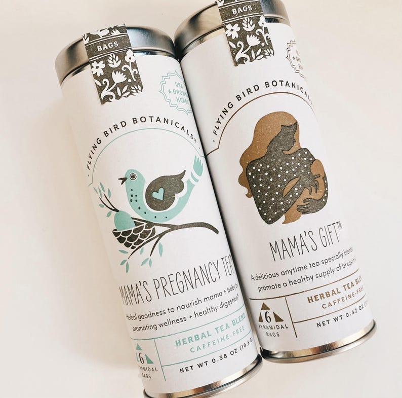 May include: Two metal tins of Flying Bird Botanicals herbal tea. The tin on the left is labeled "Mama's Pregnancy Tea" with a blue bird illustration. The tin on the right is labeled "Mama's Gift" with a brown illustration of a woman breastfeeding. Both tins have a green and brown floral design and are labeled with "6 Pyramidal Bags".
