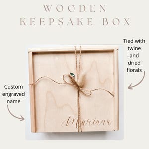 May include: A wooden keepsake box with a light wood finish. The box is tied with twine and dried florals. The name "Mariana" is custom engraved on the front. The text "WOODEN KEEPSAKE BOX" is at the top.