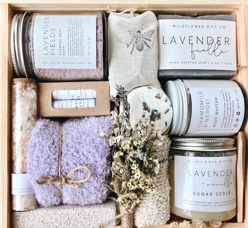 May include: A wooden gift box filled with lavender-scented bath and body products. Includes a jar of foaming bath soak, a bar of soap, sugar scrub, body butter, a loofah, and a lavender-colored washcloth. The box is labeled "Wildflower Gift Co."