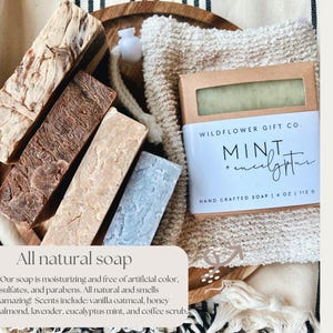 May include: An assortment of all-natural soaps in various colors and textures, including a bar labeled "Mint + Eucalyptus" by Wildflower Gift Co. The bar is 4 oz / 113 g. The image also includes a washcloth and a wooden tray.
