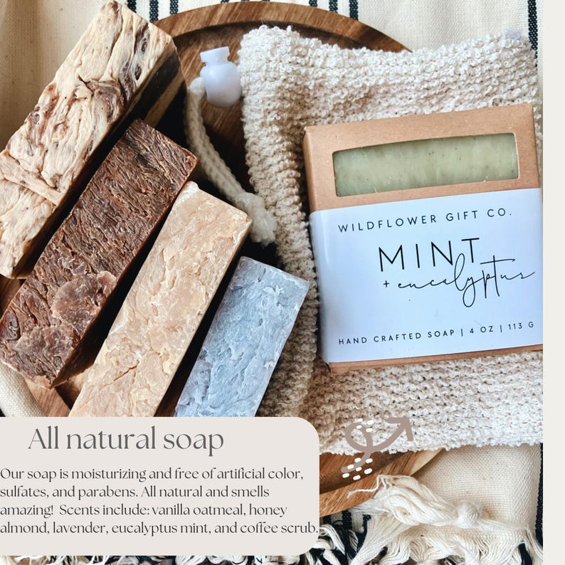 May include: An assortment of all-natural soaps in various colors and textures, including a bar labeled "Mint + Eucalyptus" by Wildflower Gift Co. The bar is 4 oz / 113 g. The image also includes a washcloth and a wooden tray.