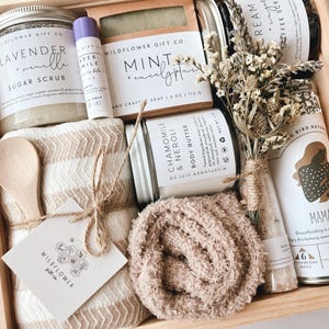 May include: A wooden gift box filled with self-care items, including a lavender sugar scrub, a mint and eucalyptus soap, a chamomile and neroli body butter, a small bottle of bath salts, a brown fuzzy blanket, a wooden spoon, and a small jar of herbal tea. The box is decorated with a small tag that says "Wildflower Gift Co."