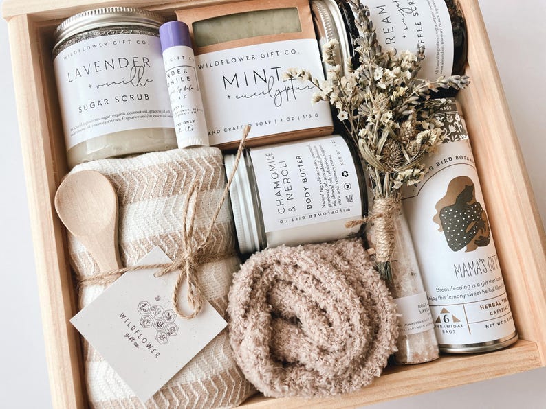 May include: A wooden gift box filled with self-care items, including a lavender sugar scrub, a mint and eucalyptus soap, a chamomile and neroli body butter, a small bottle of bath salts, a brown fuzzy blanket, a wooden spoon, and a small jar of herbal tea. The box is decorated with a small tag that says "Wildflower Gift Co."