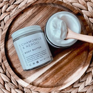 May include: A jar of Chamomile & Neroli body butter with a white lid and label, next to an open jar of body butter with a wooden spoon. The label lists natural ingredients and the net weight of 4 oz.