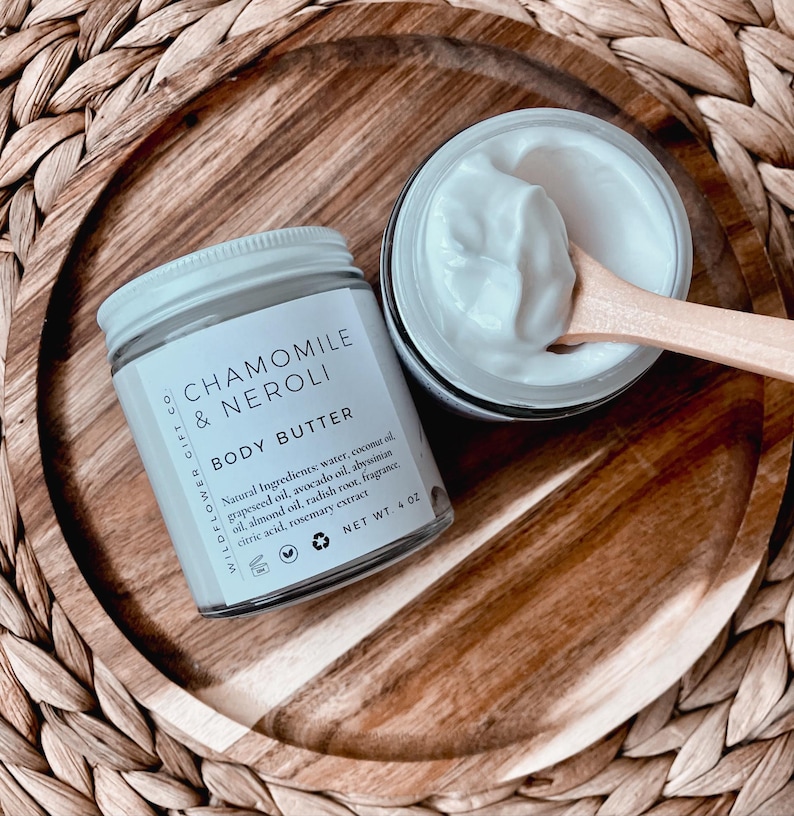 May include: A jar of Chamomile & Neroli body butter with a white lid and label, next to an open jar of body butter with a wooden spoon. The label lists natural ingredients and the net weight of 4 oz.