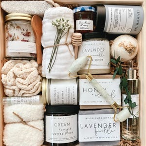 May include: A wooden gift box filled with various bath and body products, including a white bathrobe, a jar of bath salts, a bar of soap, a bottle of body butter, a jar of coffee scrub, a bath bomb, a bottle of facial oil, and a jade roller. The box is decorated with a sprig of greenery and a small wooden spoon.
