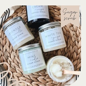 May include: An overhead shot of several jars of sugar scrub on a woven basket. The jars have silver lids and labels with text like "Lavender & Vanilla" and "White Tea + Ginger." A wooden spoon scoops out the scrub from an open jar.
