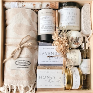 May include: A wooden gift box filled with self-care items, including a beige waffle weave towel, a jar of white tea and ginger sugar scrub, a bar of lavender fields soap, a bar of honey and almond soap, a small jar of lavender bath salts, a bottle of chamomile dreams facial oil, and a small jar of orange blossom bath salts. The box also includes dried flowers and a wooden spoon.