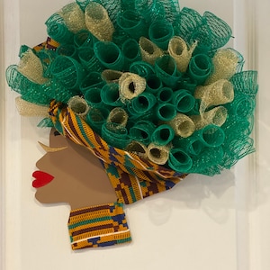 May include: A green and gold wreath with a brown silhouette of a woman's face. The woman's face is wearing a patterned head wrap and a matching collar.