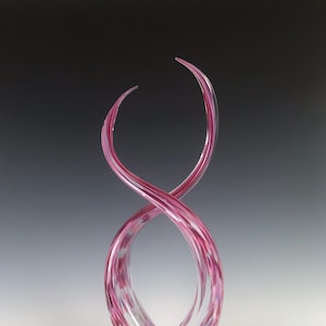 Op de afbeelding: Een roze en witte glazen sculptuur met een wervelend, abstract ontwerp. De sculptuur is hoog en slank, met een delicate, vloeiende vorm.