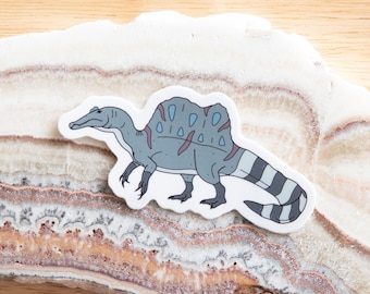 May include: A gray and white Spinosaurus dinosaur sticker with blue accents. The sticker is on a white and brown background.