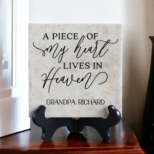 May include: A beige ceramic tile with black text that reads "A piece of my heart lives in heaven" and "Grandpa Richard". The tile is on a black stand.